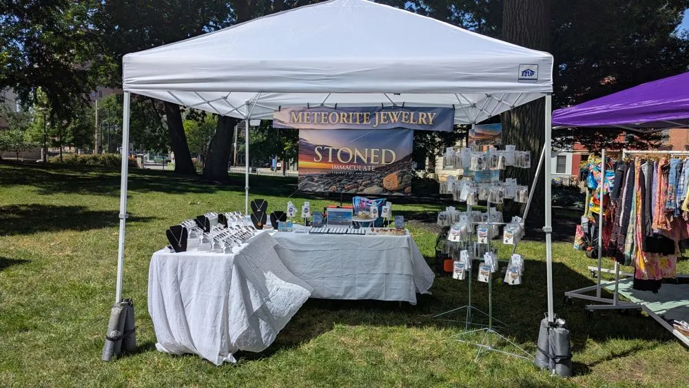 The Stoned Immaculate booth outdoors at an arts and crafts show, with tumbled stone pendants and meteorite earrings on display under a white canopy