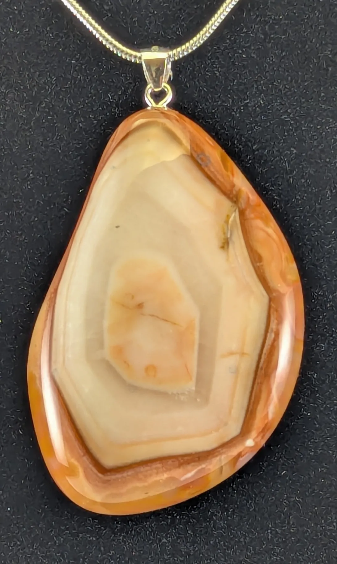 Teardrop-shaped agate pendant with a rusty orange rim, nested angular cream and tan bands, and a small peach-colored center, on a silver-tone bail and snake chain, against a black background