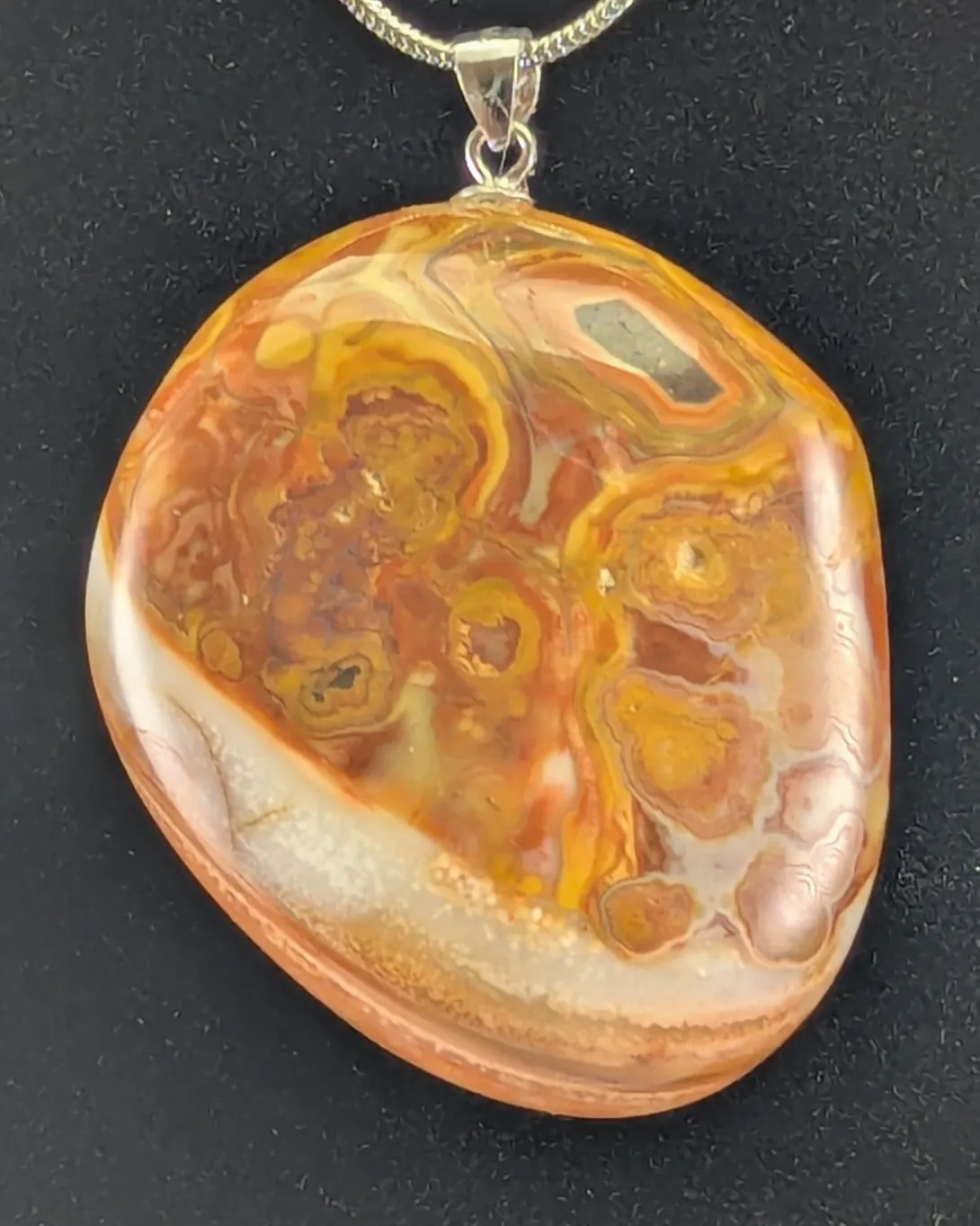 Rounded Bahia agate pendant covered in orange and gold eye formations with pale centers, with a cream band across the lower edge, on a silver-tone bail and snake chain, against a black background