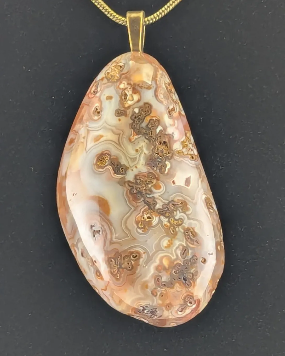 Elongated agate pendant in pale pink and cream with clusters of small rust-brown orbs and rings, on an antique bronze-tone bail and snake chain, against a black background