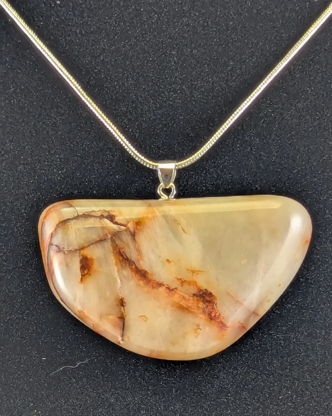 Wide, flat quartz pendant in cream and pale honey tones with thin rust-red veins running across it, on a silver-tone bail and snake chain, against a black background