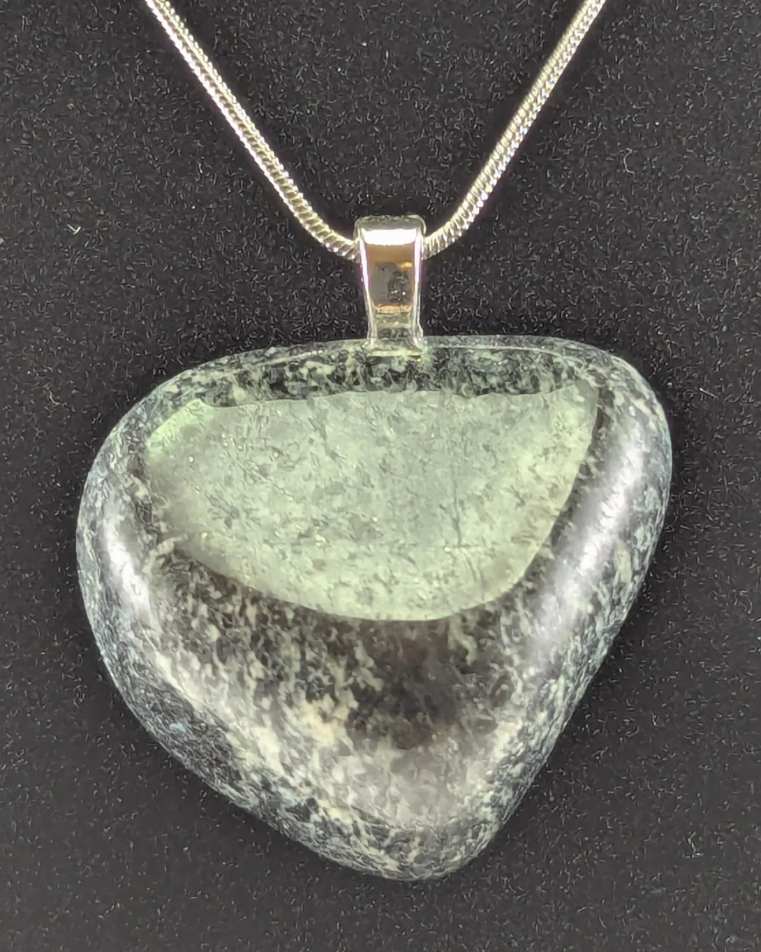 Rounded heart-shaped granite pendant speckled with dark gray and pale gray-green mineral grains, on a silver-tone bail and snake chain, against a black background
