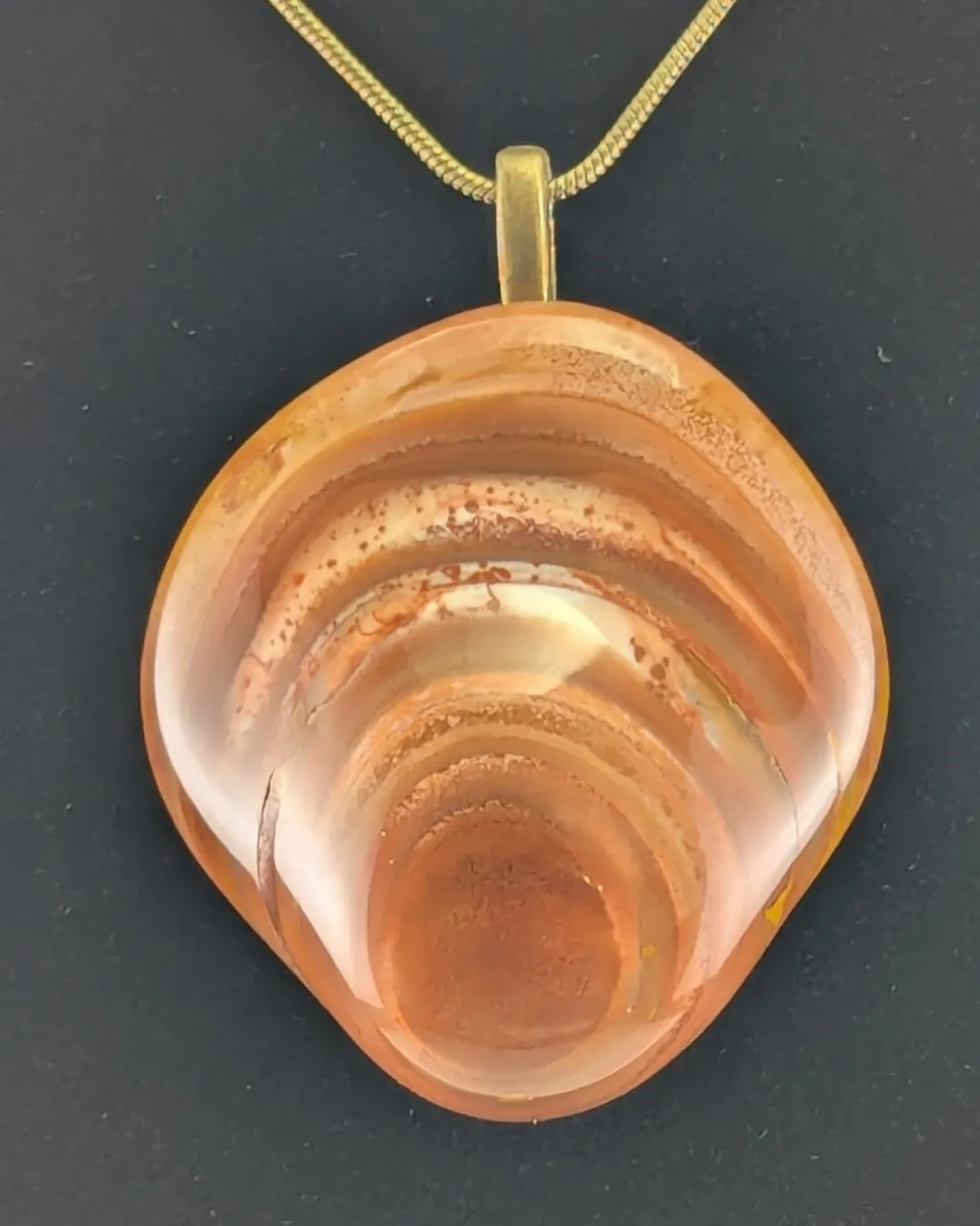 Rounded peach and pale orange Bahia agate pendant with stacked concentric rings that resemble a seashell, on an antique bronze-tone bail and snake chain, against a black background