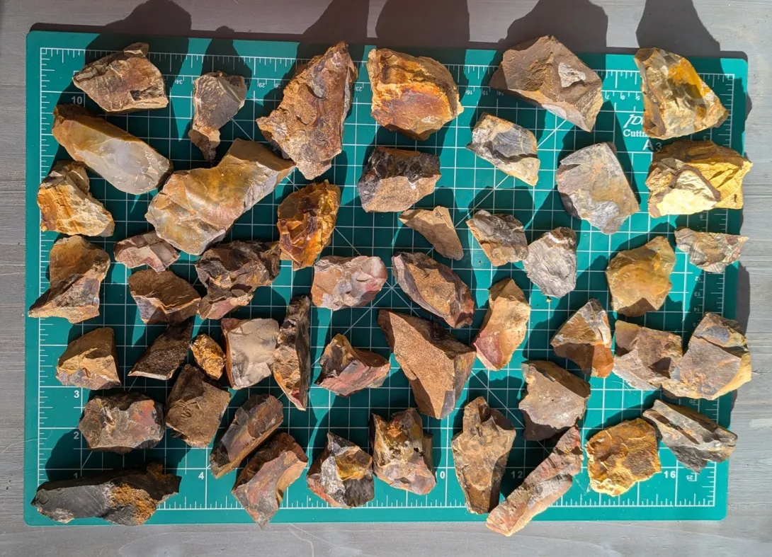Dozens of raw, broken pieces of Ohio Flint Ridge flint in rust, gold, tan, grey and reddish tones, with sharp angular edges, spread out on a green cutting mat with ruler markings