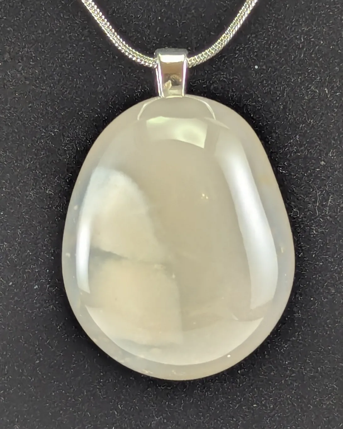 Large oval pendant of pale, milky chalcedony with soft cloudy patches inside, on a silver-tone bail and silver chain against black velvet