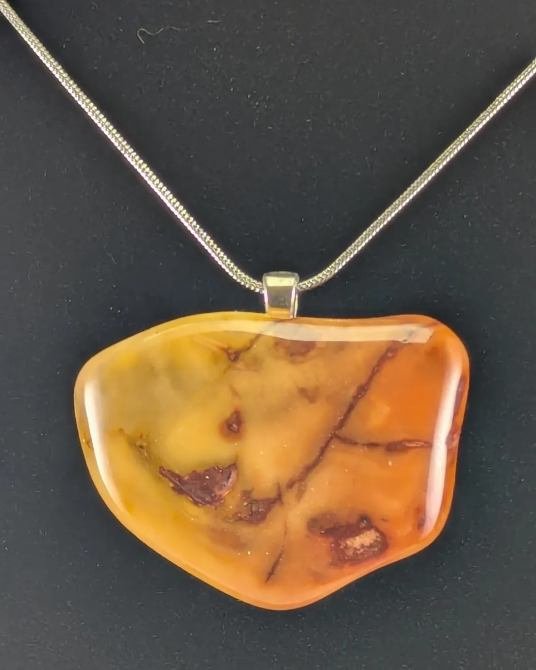 Flat, irregular jasper pendant shading from honey yellow to deep orange, with thin dark red veins branching across it, on a silver-tone bail and snake chain, against a black background